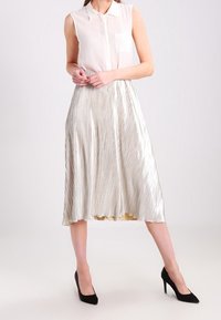 Blouse sans manches blanche légère et transparente avec une poche. Jupe midi plissée argent métallique, longueur mi-mollet, avec une texture lisse, associée à des talons noirs.