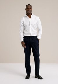 Biała koszula na długi rękaw z guzikami, ciemne spodnie i czarne buty. Koszula ma kołnierzyk i dopasowany krój, z gładką fakturą.