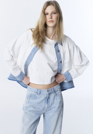 Jonge vrouw met lang blond haar, gekleed in een witte cropped top, een witte en denim jas, en lichtblauwe jeans met steentjes.