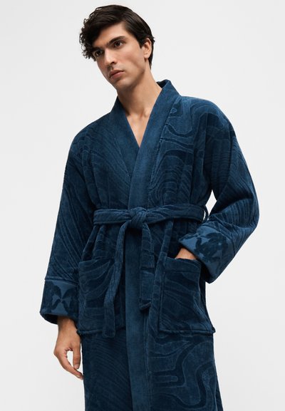 Homme portant un peignoir bleu foncé texturé avec une ceinture nouée et de grandes poches avant sur un fond clair uni.