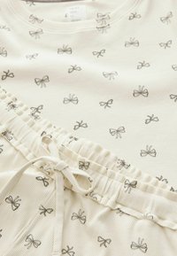 Light cream loungewear set featuring a top and bottoms with a repeating bow pattern. Ribbed texture, elastic waistband, and drawstring detail.