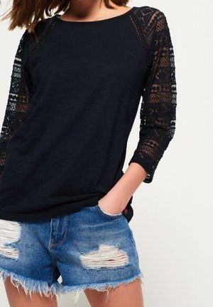 Vrouw draagt een zwart shirt met lange mouwen van kant en blauwe denim shorts met rafelige zomen, hand in de zak gestopt.