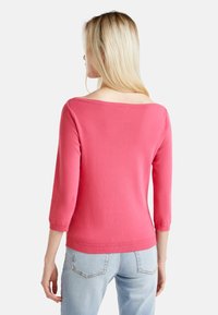 Maglione rosa lavorato a maglia con maniche a tre quarti, scollatura ampia e polsini e orlo a costine. Indossato con jeans azzurri chiari.