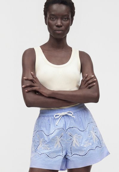 Femme les bras croisés portant un haut sans manches crème et un short bleu clair avec une broderie blanche de palmier et une taille à cordon.