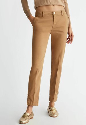 Pantaloni in cotone beige con vestibilità slim, tasche anteriori e un piccolo dettaglio con zip alla caviglia, abbinati a mocassini dorati.