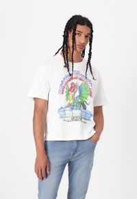 White cotton t-shirt featuring a colorful dragon graphic and text, paired with light blue denim jeans. The fit is relaxed.