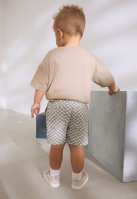 Chemise beige à manches courtes, short à carreaux gris, chaussettes blanches et baskets claires. Debout contre un cube bleu dans un espace minimaliste.