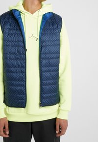 Niebieska pikowana kamizelka z geometrycznym wzorem, z zamkiem błyskawicznym i kontrastującą niebieską podszewką, noszona na neonowozielonej bluzie z kapturem.