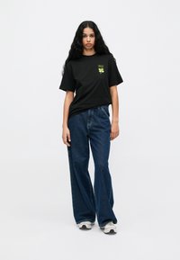 Must black t-särk lühikeste varrukatega ja rohelise graafilise disainiga, kombineeritud laia jalgadega siniste teksade ja valgete tossudega.