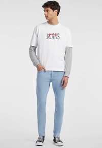 Camiseta blanca con el logo de "Guess Jeans" en rojo y gris, superpuesta con una camisa de manga larga gris, combinada con jeans de mezclilla azul claro y zapatillas negras.