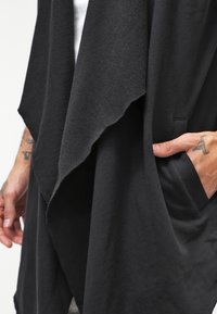 Hand med tatueringar som vilar i fickan på ett svart draperat plagg med asymmetrisk lager av tyg.