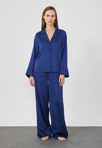 Set di pigiama in satin blu navy composto da una camicia con bottoni a maniche lunghe e pantaloni a gamba larga, decorato con un motivo di stelle e lune crescenti.