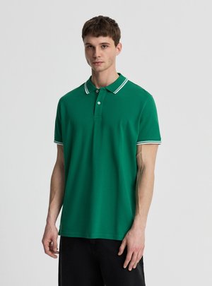 Młody mężczyzna w zielonej koszulce polo z krótkim rękawem, z białym paskowanym kołnierzykiem i mankietami, stojący na jednolitym jasnoszarym tle.