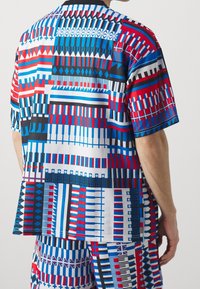 Camisa de manga corta con un atrevido patrón geométrico en azul, rojo y negro sobre un fondo blanco, hecha de una tela ligera con un ajuste relajado.