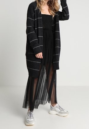 Cardigan nero con strisce bianche, indossato sopra una gonna in mesh nera. Abbinato a sneaker chiare. Tessuto morbido, design aperto davanti.