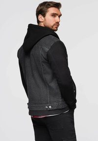 Felpa nera abbinata a una giacca di jeans scura. Presenta texture a contrasto, bottoni a pressione in vita e un look a strati con una camicia a quadri sotto.