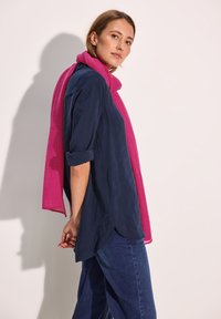 Mørkeblå bluse med opsmøgede ærmer og længere ryg. Viklet i et ribbet lyserødt tørklæde. Sammen med løstsiddende denimjeans.