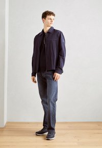 Veste légère marine avec une fermeture éclair et un col pointu, associée à un jean bleu foncé et des baskets grises sur un sol en bois.