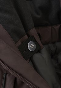 Brunt tyg med en mjuk textur har en knapp fäst på ett svart elastiskt band. Delvis synlig grå foder med sömnadsdetalj.