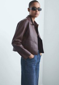 Brun lädercroppad jacka med krage och två framfickor, kombinerad med högmidjade blå denimjeans. Modellen bär svarta solglasögon.