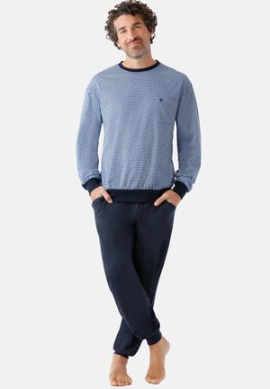 Man standing barefoot with hands in pockets wearing a light blue patterned long-sleeve top and dark blue pants against a plain background.
