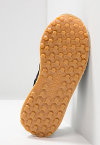 Musta lenkkarit, joissa on teksturoitu kumipohja, hunajakenno-kuvio ja vaaleanruskea väri, kohokuvioidulla logolla ja kaarevalla siluetilla.