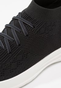 Zwarte gebreide sneaker met een sokachtige ontwerp, voorzien van platte donkergrijze veters, een gestructureerd bovenwerk en een witte rubberen zool.