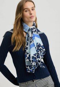 Foulard in seta con una stampa leopardata blu e bianca, caratterizzato da diverse sfumature di blu. Indossato sopra un maglione a coste blu navy con dettagli nei bottoni.