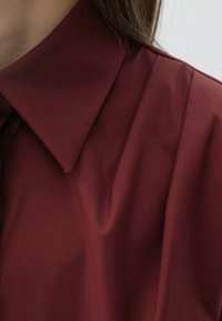 Bordowa koszula z usztywnionym kołnierzykiem, wykonana z gładkiego, lekko pomarszczonego materiału, który podkreśla jej układ i fakturę.