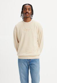 Beige sweatshirt med preget "Levi's" logo, rund hals, vide ermer og ribbestrikkede mansjetter. Kombinert med blå denimjeans.