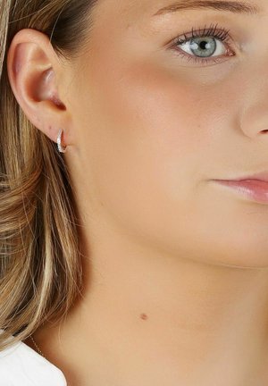 Silverfärgade hoopsmycken med en slät, böjd design och små infällda klara ädelstenar. Bärs på en models öra med mellanhår av blont hår.