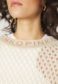 Pull en maille blanche avec un tissage ouvert texturé, tissu couleur crème et un col beige portant "PINKO" en relief rose clair. Boucles d'oreilles statement dorées.