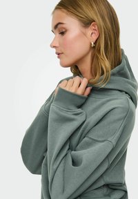 Groene katoenen hoodie met een grote capuchon en geribbelde mouwen, gedragen door een model. Heeft een relaxte pasvorm en een zachte textuur.