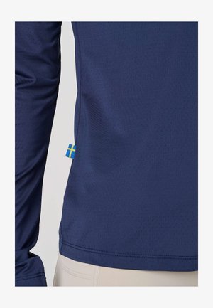 Marineblauw geperforeerd hemd met lange mouwen en een klein Zweeds vlaggetje op de onderste zijnaad, gedragen over lichtbeige broek.