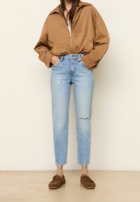 Mujer vistiendo una chaqueta marrón con cremallera, jeans desgastados de color azul claro y zapatos mocasines marrones, de pie contra un fondo plano.