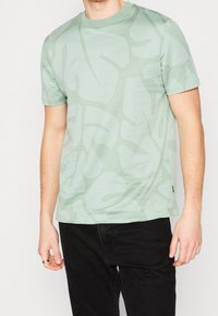 Vaaleanvihreä lyhythihainen t-paita, jossa on teksturoitu lehtikuvio. Pyöreä pääntie ja suora leikkaus; yhdistettynä tummiin farkkuihin.