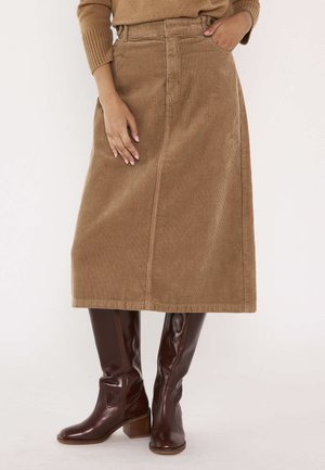 Donna con gonna in velluto a coste beige al ginocchio, maglione abbinato e stivali al ginocchio in pelle marrone scuro, in piedi con una mano in tasca.