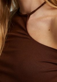 Ruskea ribattu tankkitoppi, jossa on leveä pääntie ja kapeat olkaimet. Kangas vaikuttaa pehmeältä ja siinä on hienovarainen tekstuuri.