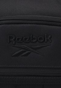 Fekete Reebok táska textúrált anyagból, dombornyomott logóval és kontrasztos varrással a cipzárnál. Egyszerű, funkcionális dizájn.