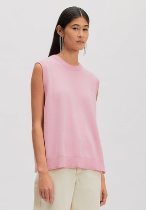Vrouw die een mouwloos, loszittend roze gebreid topje en crèmekleurige broek draagt, geaccentueerd met lange, hangende strass oorbellen.