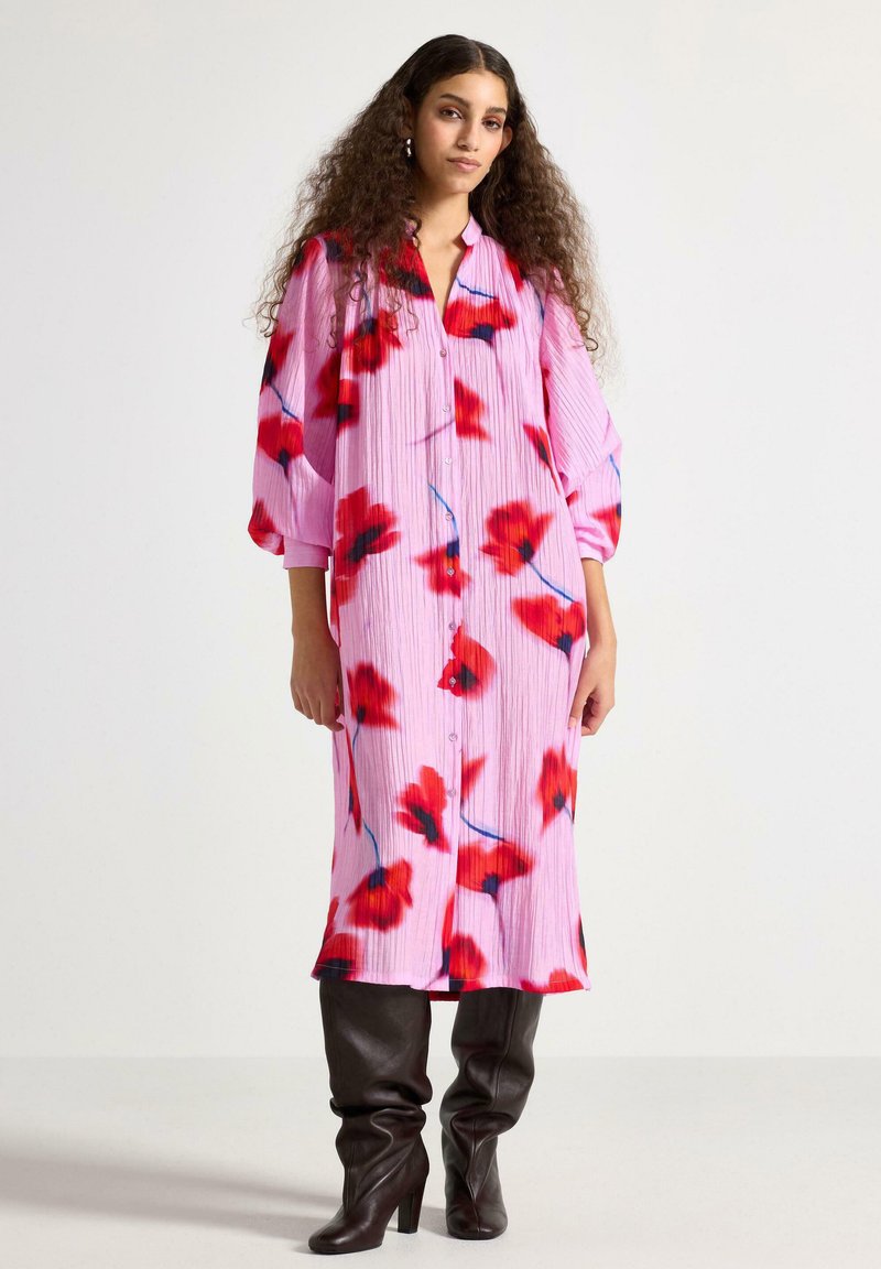 Lang lyserød kjole med rødt blomsterprint, med knappelukning foran, plisseret tekstur og løse ærmer, kombineret med mørke knælange støvler.