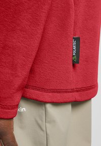 Rode fleecetrui met een gladde textuur, voorzien van een Polartec-label. Het onderste deel toont een lichtbeige stof, met een zichtbare naad en stikseldetails.