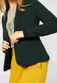 Mörkgrön kavaj med en mjuk textur, slim fit, långa ärmar och en enkel knappstängning. Kombineras med en grå grafisk t-shirt och gula byxor.