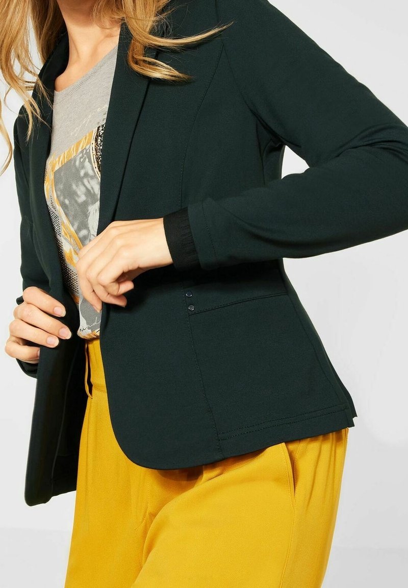 Mörkgrön kavaj med en mjuk textur, slim fit, långa ärmar och en enkel knappstängning. Kombineras med en grå grafisk t-shirt och gula byxor.