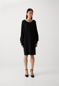 Black oversized sweater dress with long sleeves, round neckline, soft texture, and hemline falling above the knee. Worn with black heels.
