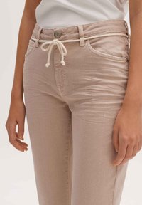 Pantalones beige claros hechos de tela texturizada con cintura de cordón, que cuentan con bolsillos frontales y cierre de botón metálico.