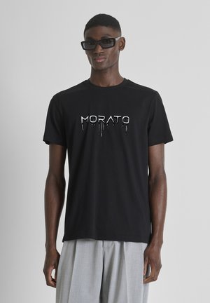 Hombre con camiseta negra con el logo "MORATO", pantalones grises plisados y gafas de sol negras rectangulares sobre un fondo gris claro.