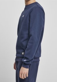 Persona vestida con un sudadera azul marino y pantalones a juego, visible desde el cuello hasta las caderas, de pie de lado contra un fondo liso.