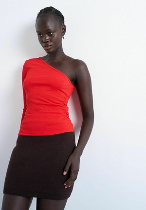 Vrouw die een rood een-schouder lang mouw top draagt en een donkerbruine nauwsluitende rok, staand tegen een effen witte achtergrond.