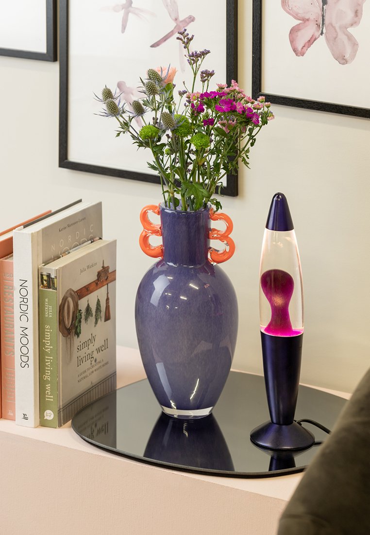Jarrón de cerámica morado con asas naranjas sosteniendo flores, al lado de libros y una lámpara de lava con una base rosa y clara sobre una superficie reflectante.